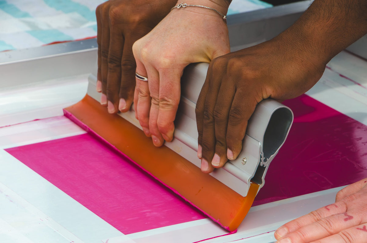 Two hands pulling a squeegee across a screen, pink ink