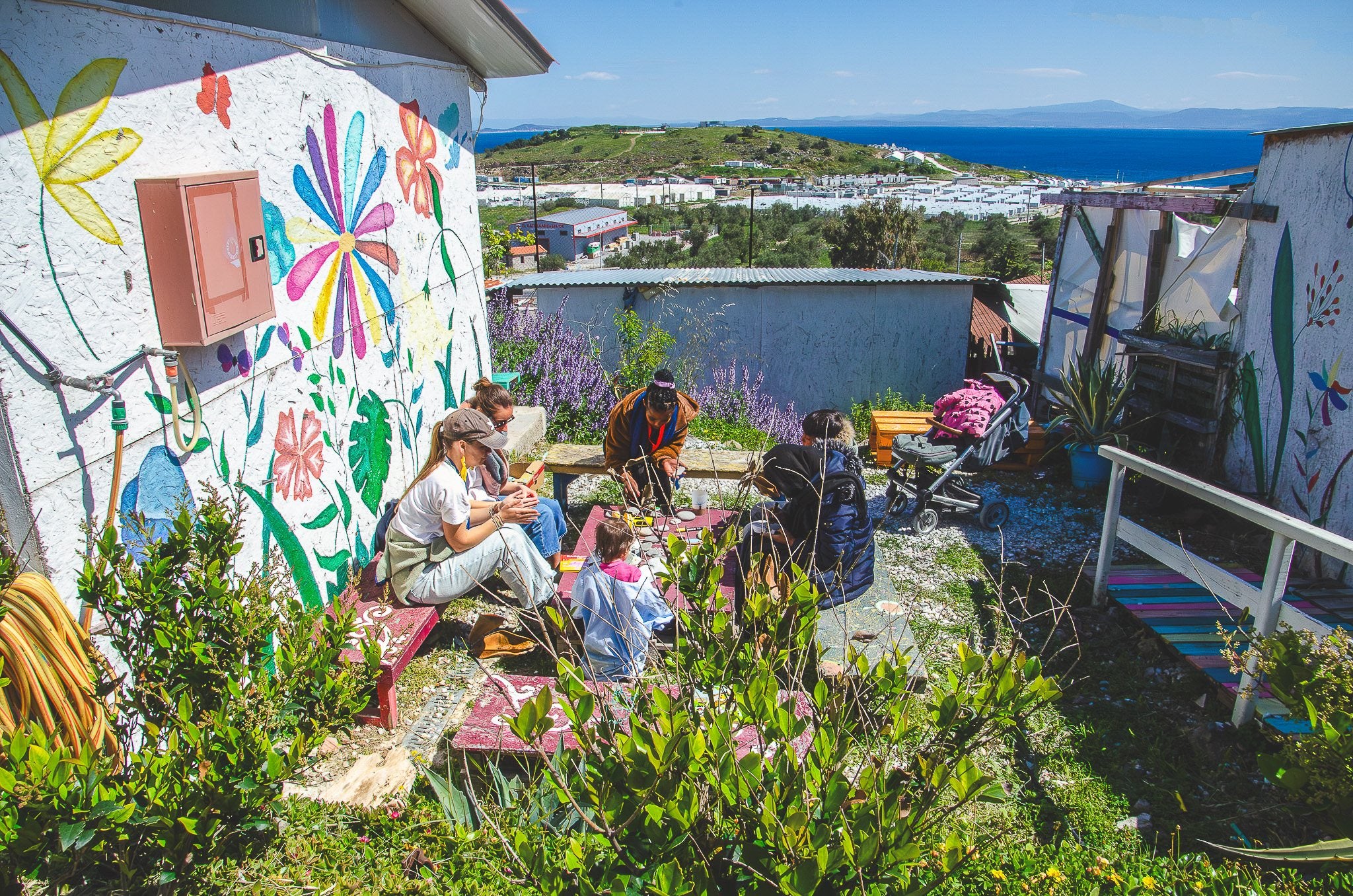 A craft table in a flowering garden at Paréa Lesvos, the sea beyond